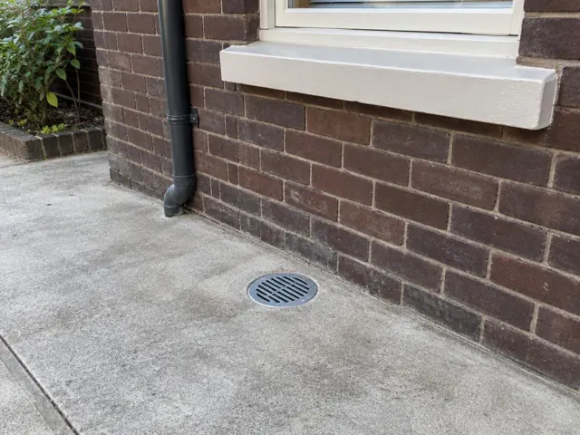 Outdoor gully grate on dry concrete beside a brick house wall in Double Bay